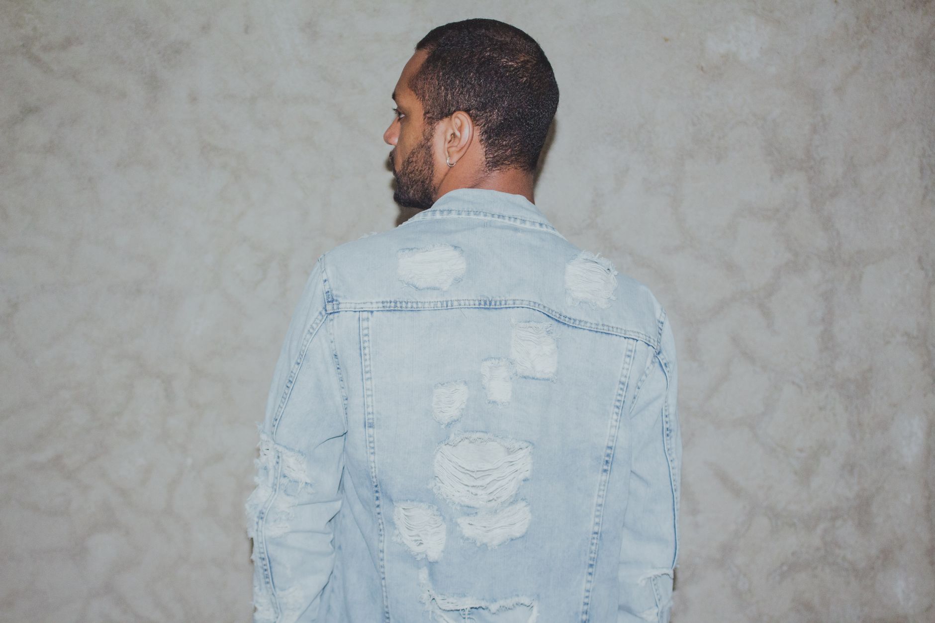 A stylish man in a tattered denim jacket poses against a textured wall.