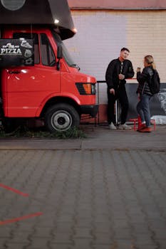 A couple enjoying drinks near a vibrant red pizza truck on a city street.