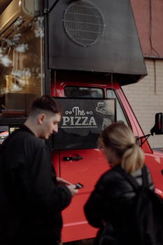 Young couple ordering pizza from a vibrant red food truck on an urban street.