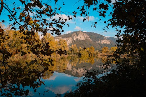 Picturesque scenery of calm lake reflecting mountains and trees with autumnal foliage in sunny day with blue cloudy sky