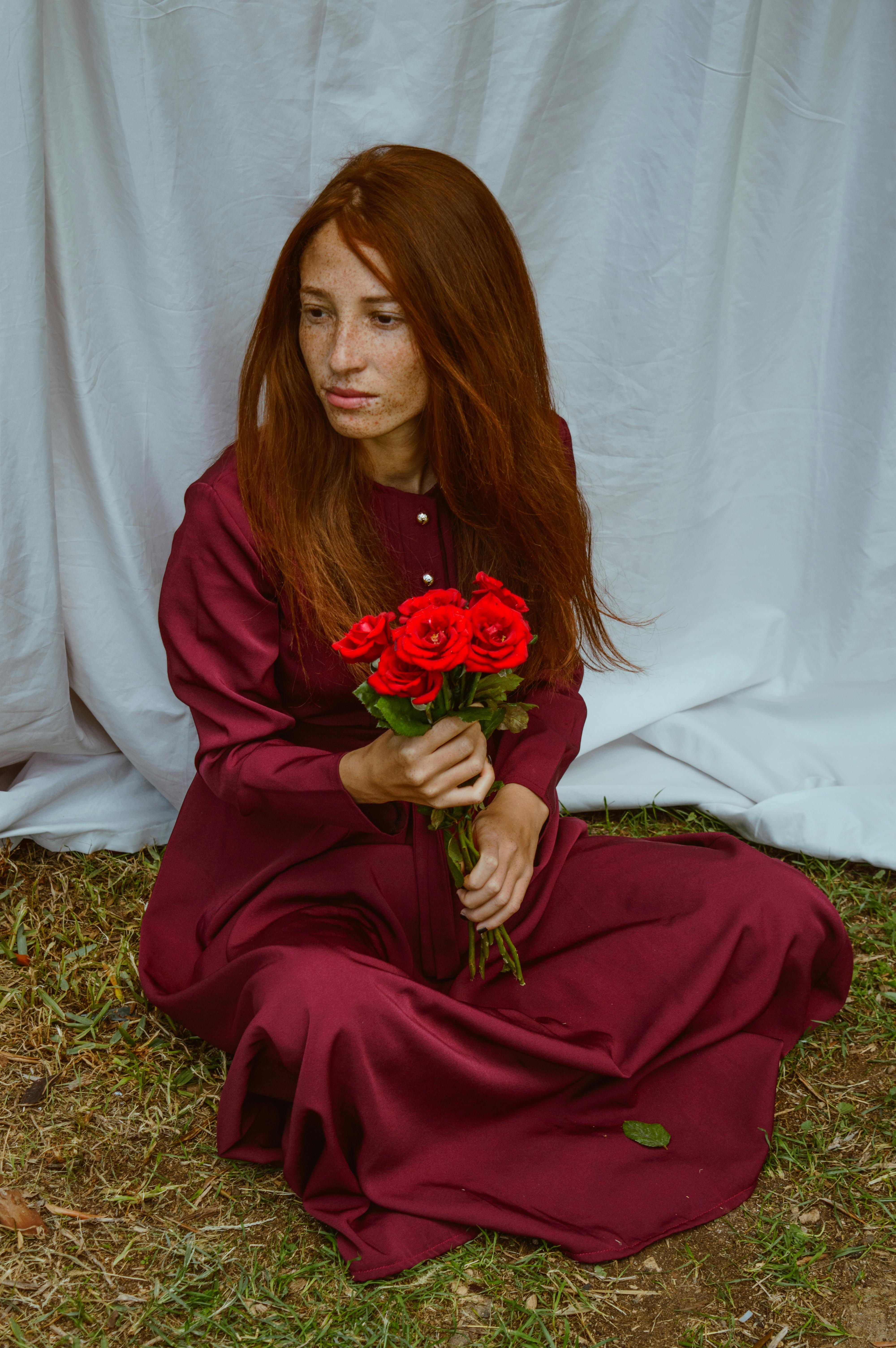 From above of tranquil young lady with long red hair in elegant maxi dress sitting on grassy ground with crossed legs with bunch of red roses in hand