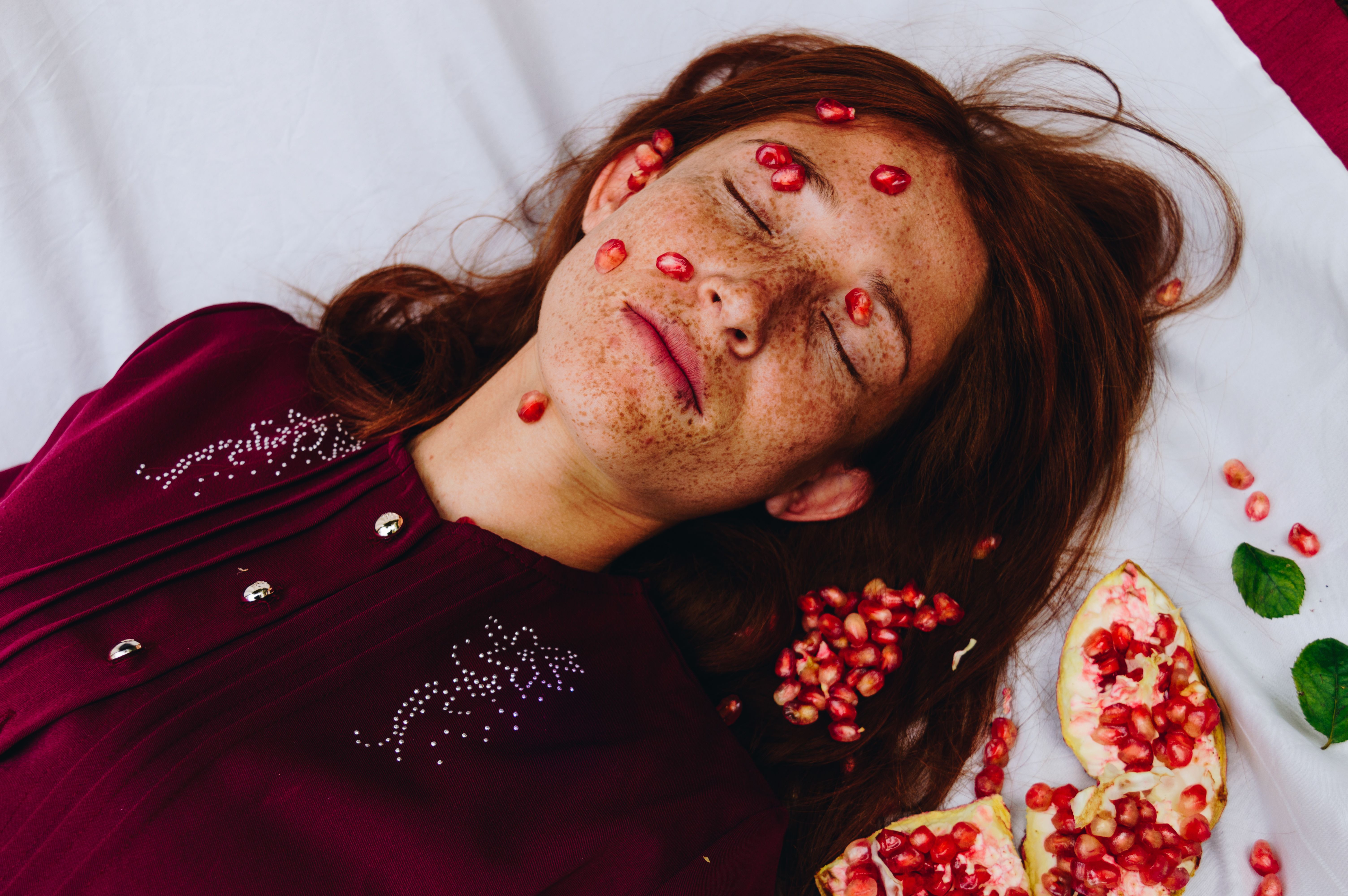 From above of young calm female with long red hair and freckles lying on comfortable bed with closed eyes and pomegranate grains on face