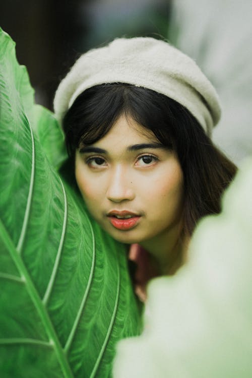 Pensive young ethnic female with dark hair in white beret looking at camera with mouth opened near big verdant leaf of exotic plant in daytime