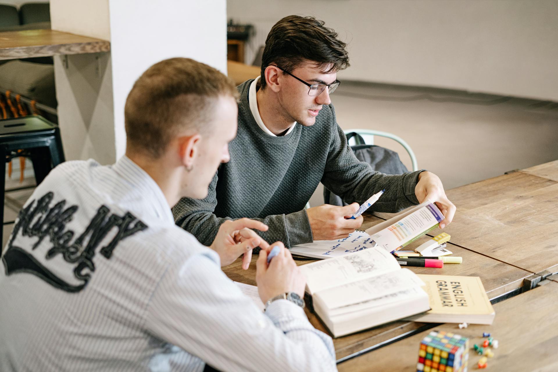 Two young men studying together, focusing on English grammar books in a modern indoor setting.