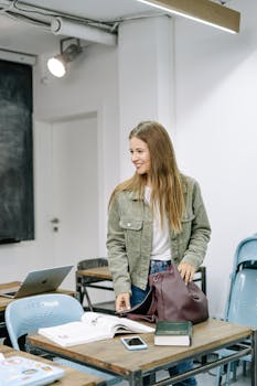 Young woman in a classroom, smiling and standing near a desk with a backpack.