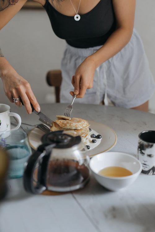 Free Woman cutting pancakes with blueberries, accompanied by coffee, in a cozy kitchen setting. Stock Photo
