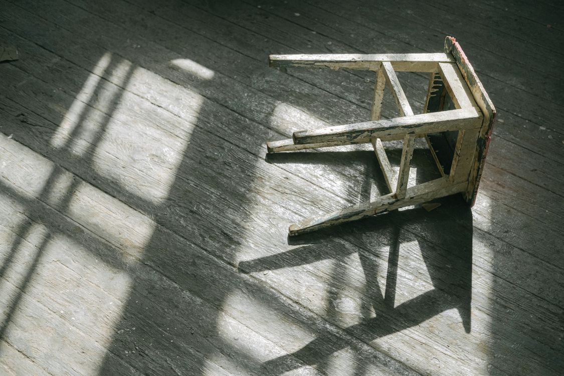 A worn wooden stool casting shadows on a sunlit hardwood floor, showing visible wear and aging