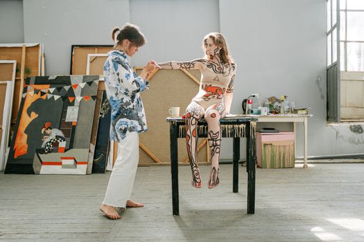 Artist painting intricate body art on model in sunlit studio.