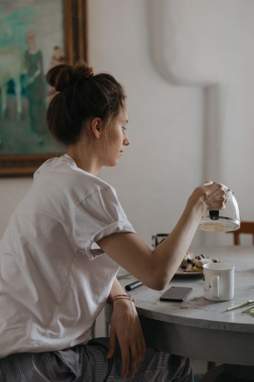 Free A young woman pours coffee into a cup at a dining table, surrounded by a cozy indoor setting. Stock Photo