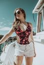 Smiling young female in sunglasses wearing colorful swimsuit and shorts standing on speedboat deck and looking away on summer day