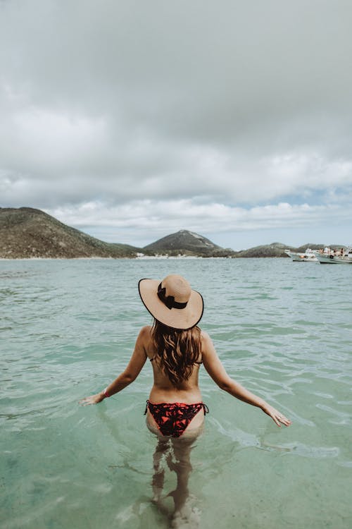 Back view of anonymous woman in swimsuit and straw hat walking in clear sea on cloudy summer day