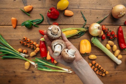 Hand holding mushrooms among an assortment of colorful vegetables on a wooden table.