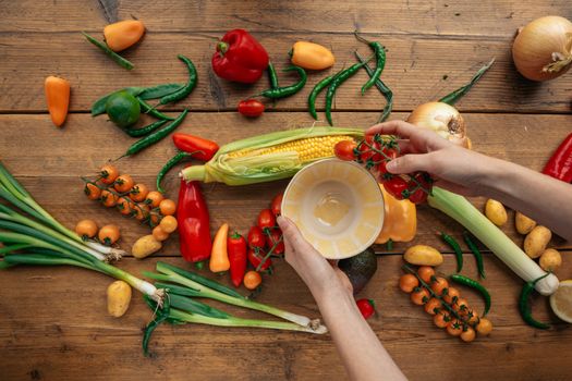 Colorful vegetables including corn, tomatoes, and peppers arranged with hands holding tomatoes in a bowl.