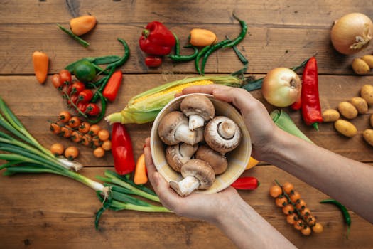 Hands hold a bowl of fresh mushrooms surrounded by vibrant vegetables on a wooden table.