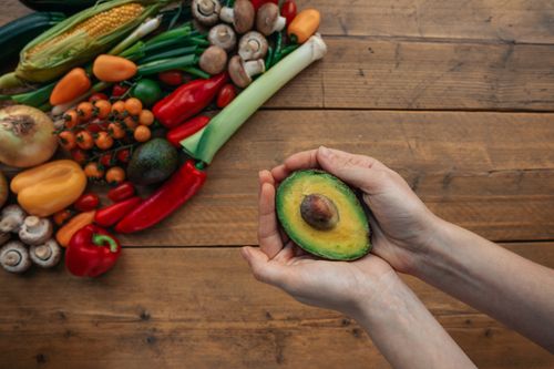 Free Hands holding a sliced avocado on a wooden table surrounded by fresh vegetables, emphasizing healthy eating. Stock Photo