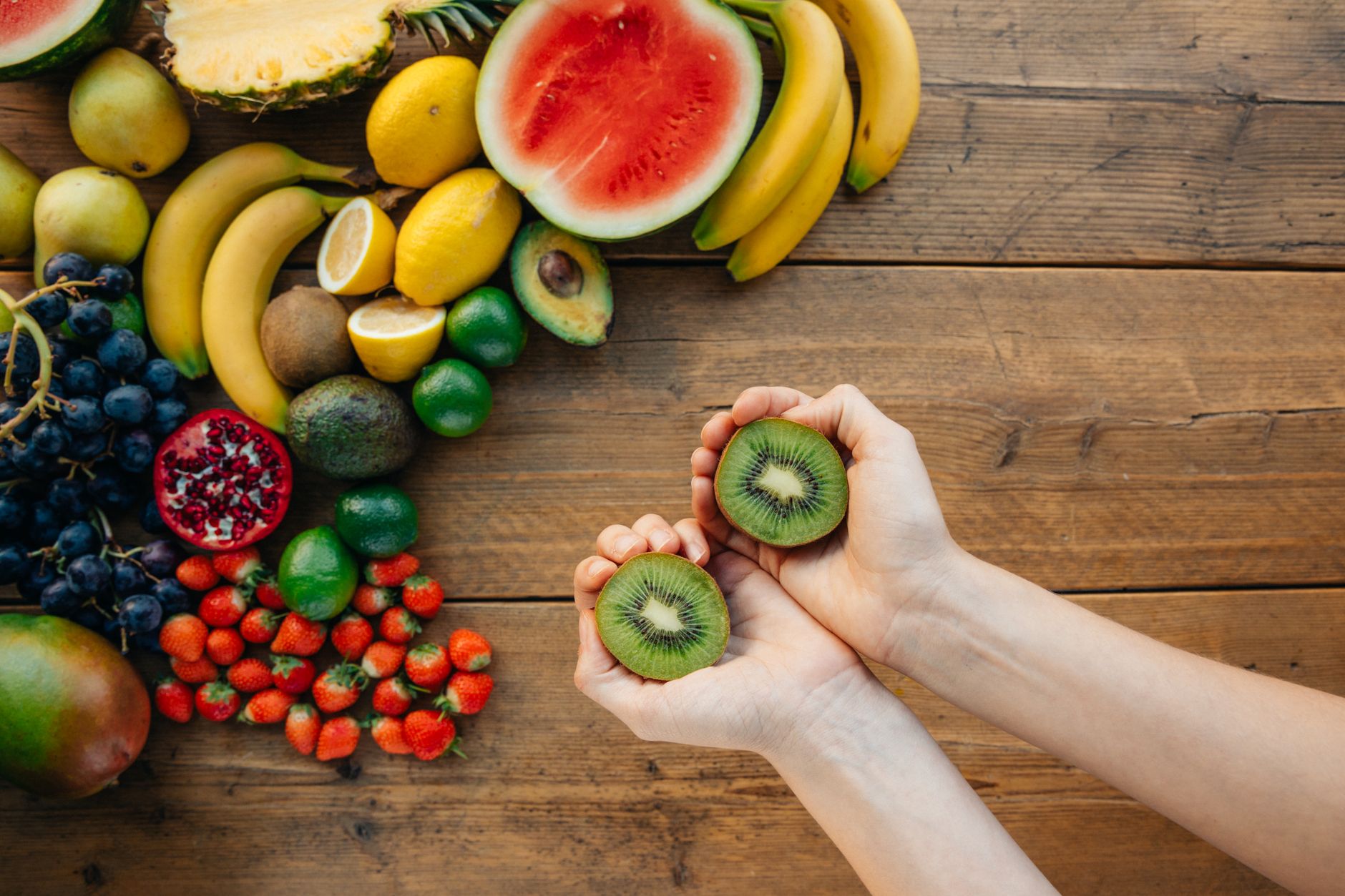 Hands holding kiwis with a variety of fresh fruits on a wooden table.
