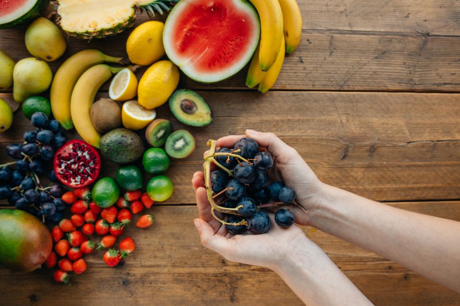 A variety of fresh, organic fruits with hands holding grapes on a wooden table.