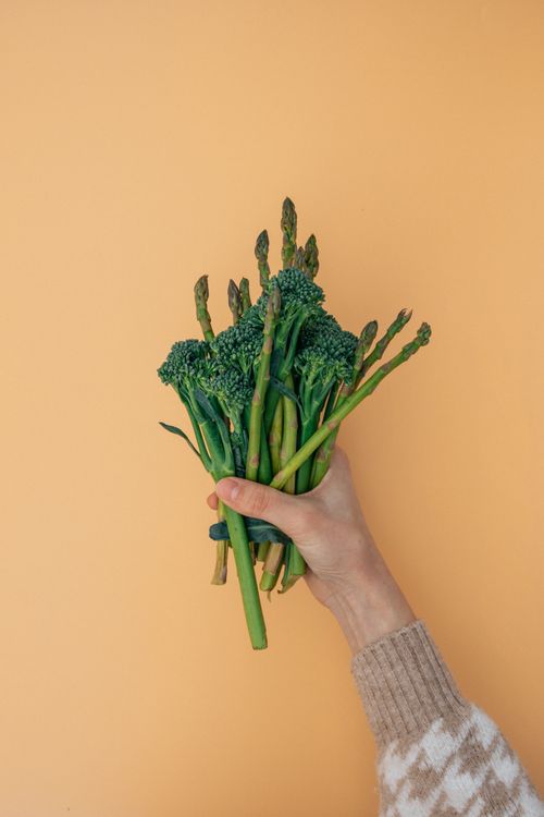 Free Hand holding fresh asparagus and broccoli against a warm orange backdrop. Ideal for healthy food themes. Stock Photo