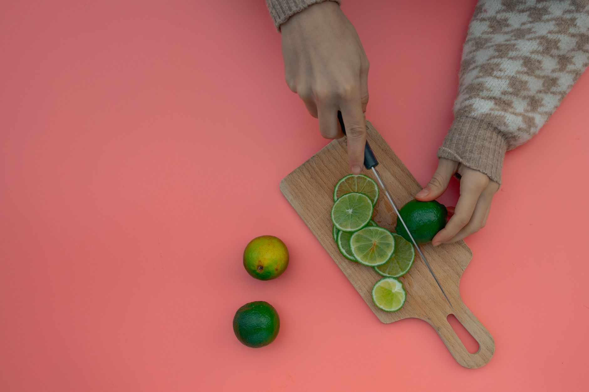 Lime benefits — Hands slicing fresh limes on a wooden chopping board against a pink background.