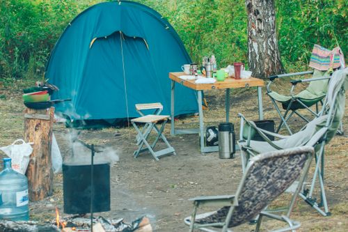 Free Serene campsite featuring a tent, chairs, and campfire amidst lush greenery for a perfect outdoor adventure. Stock Photo