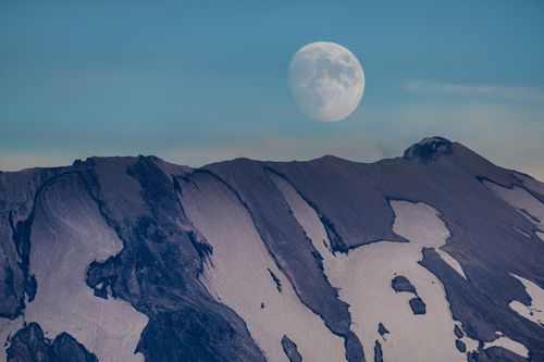 Free Beautiful moonrise over a rugged, snow-capped mountain under a clear blue sky. Stock Photo