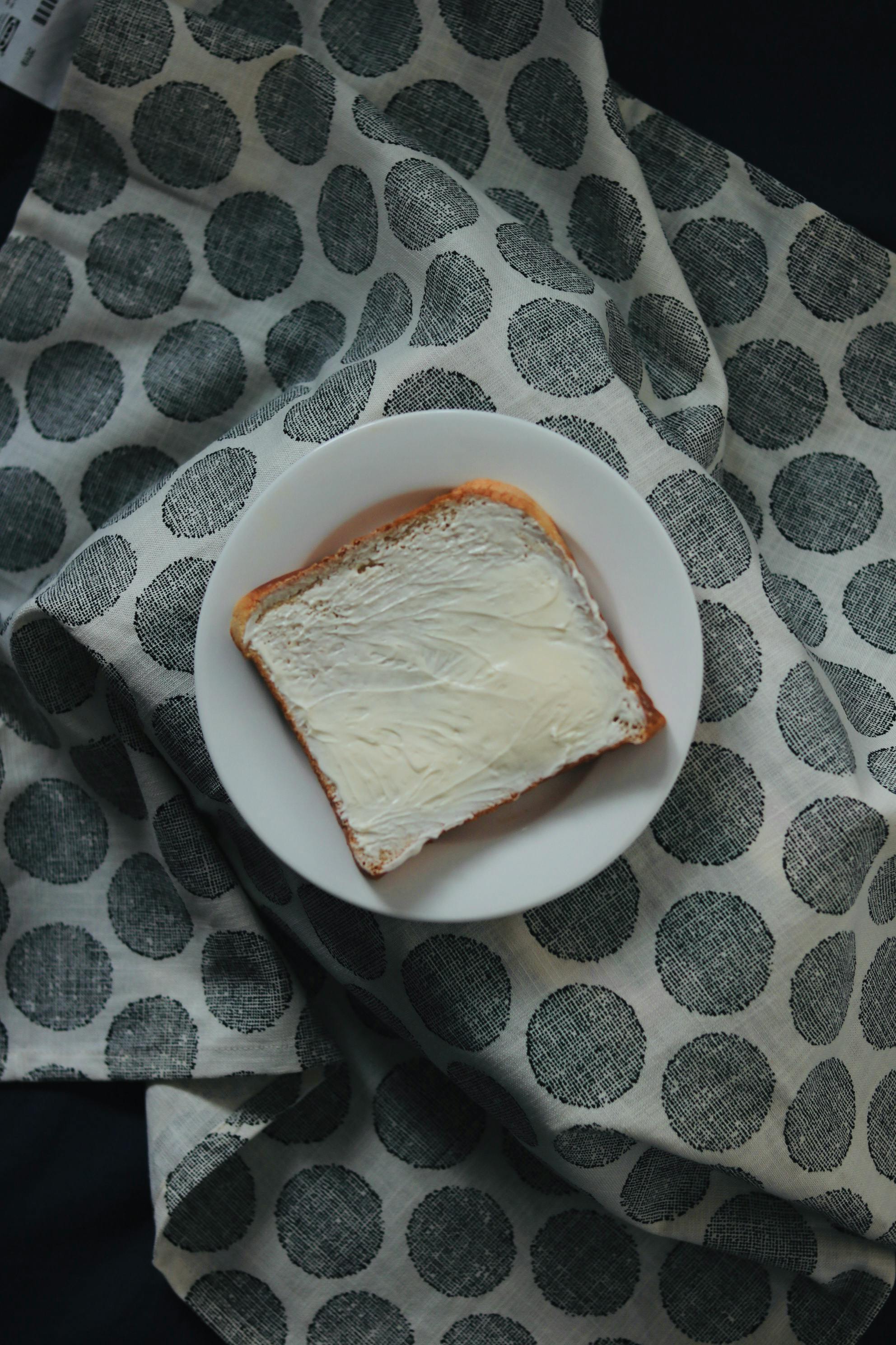 A slice of bread with butter on a white plate over a dotted cloth background.