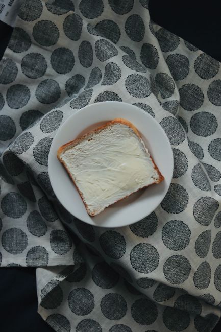 A slice of bread with butter on a white plate over a dotted cloth background.