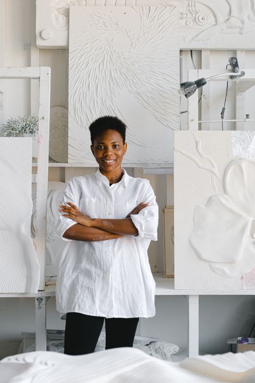 Free Portrait of a smiling African American woman artist standing in her studio surrounded by canvases. Stock Photo