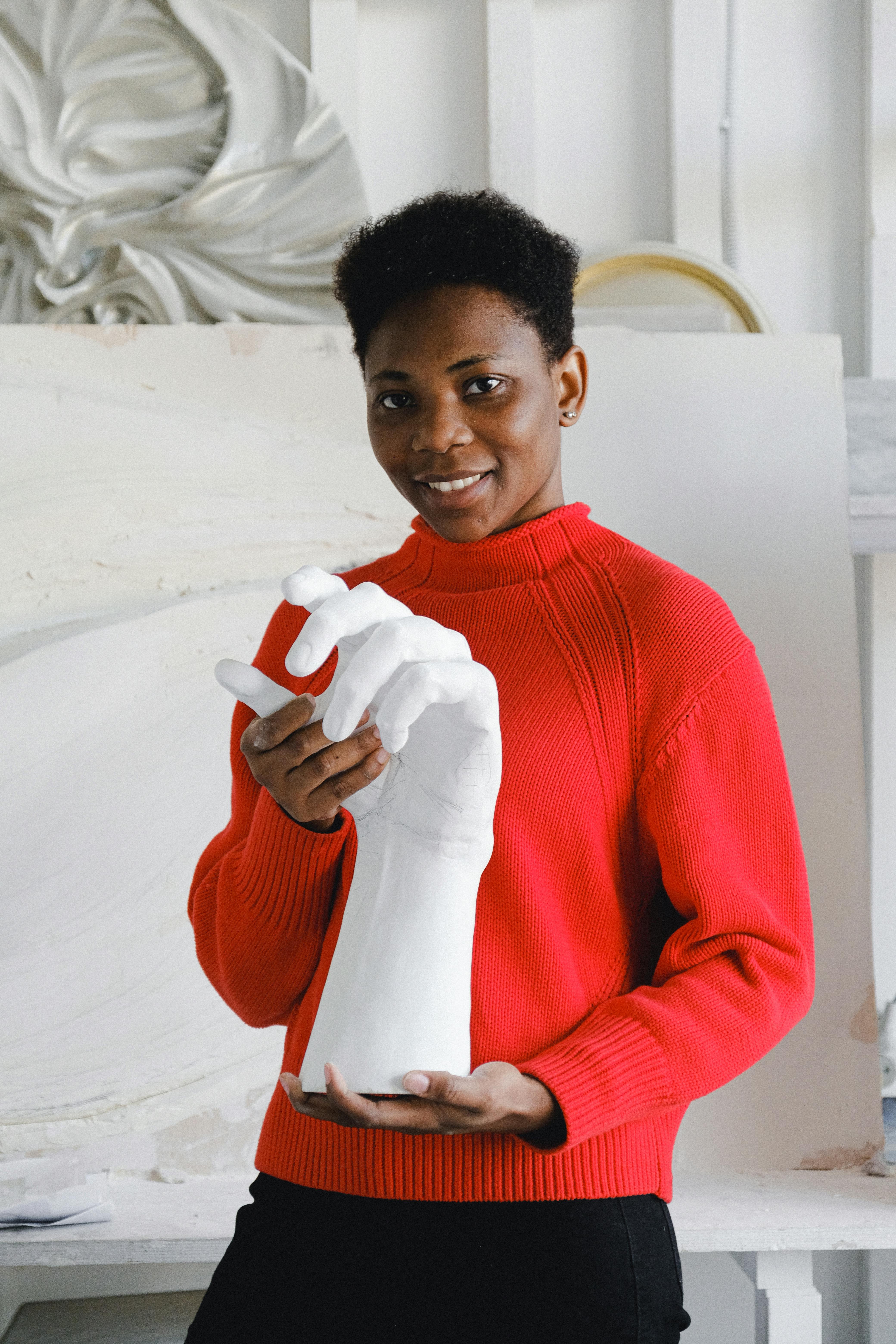 Portrait of a smiling artist in red sweater holding a white hand sculpture in an art studio.