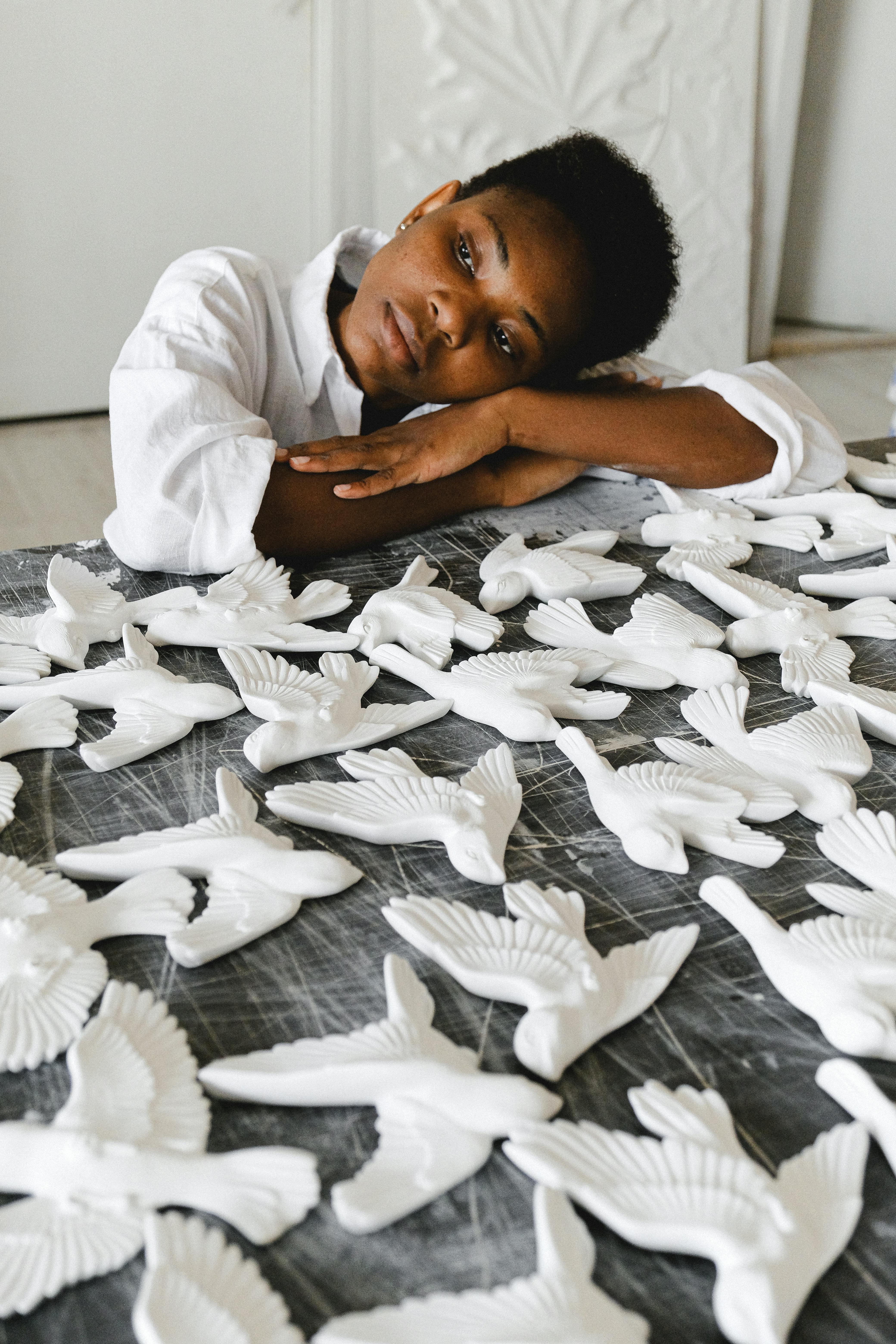 A thoughtful artist leans on a table filled with handmade ceramic birds in an indoor workshop.
