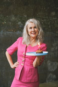 A joyful senior woman in a pink dress carrying a lit birthday cake in an indoor setting.