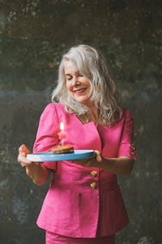 Elegant senior woman in pink holding a birthday cake with a lit candle, smiling indoors.