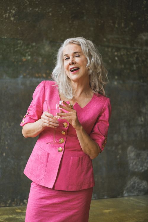 Free Cheerful senior woman in pink outfit holding a glass of wine, indoors setting. Stock Photo