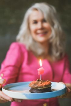 Smiling senior woman holds a small cake with a lit candle on a plate, conveying celebration.
