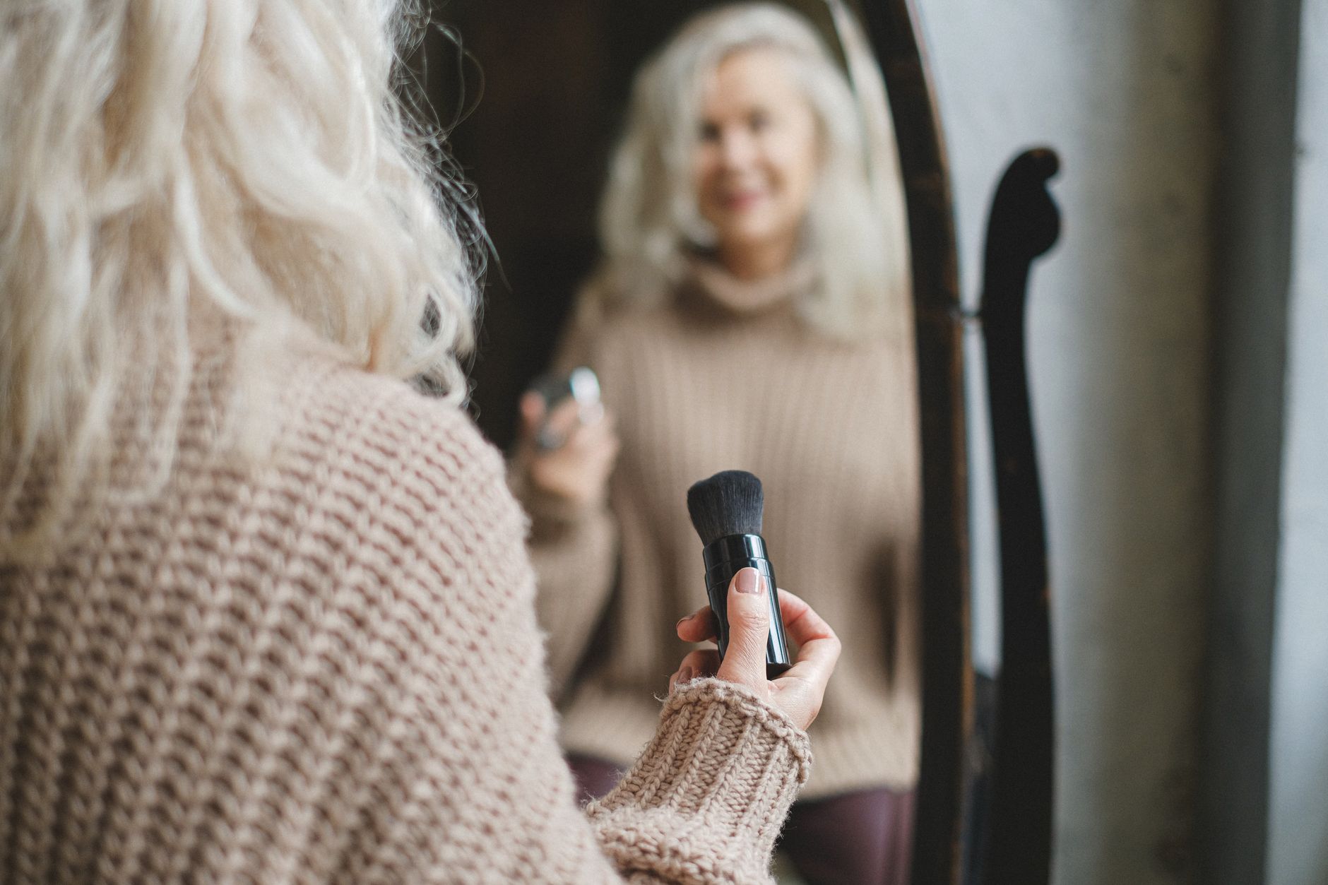 A senior woman applies makeup while looking in a mirror, holding a brush in her hand.