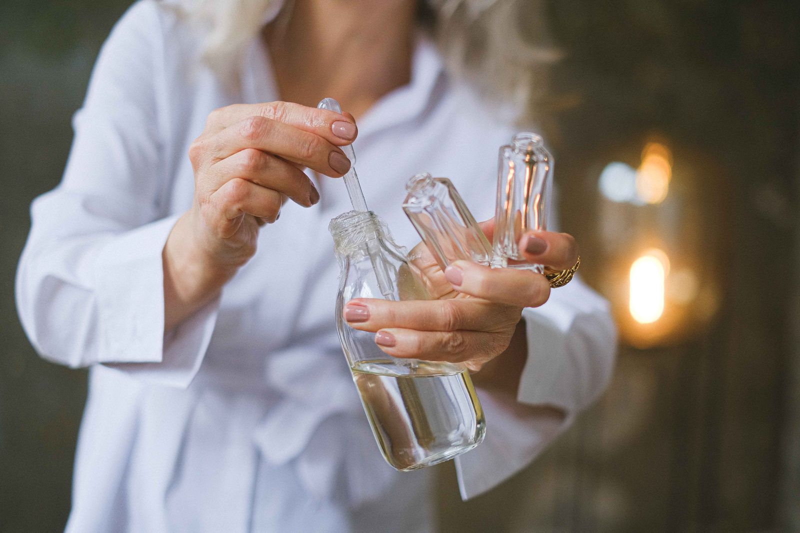 Woman in a white shirt holding a glass dropper and skincare bottles in warm ambient light