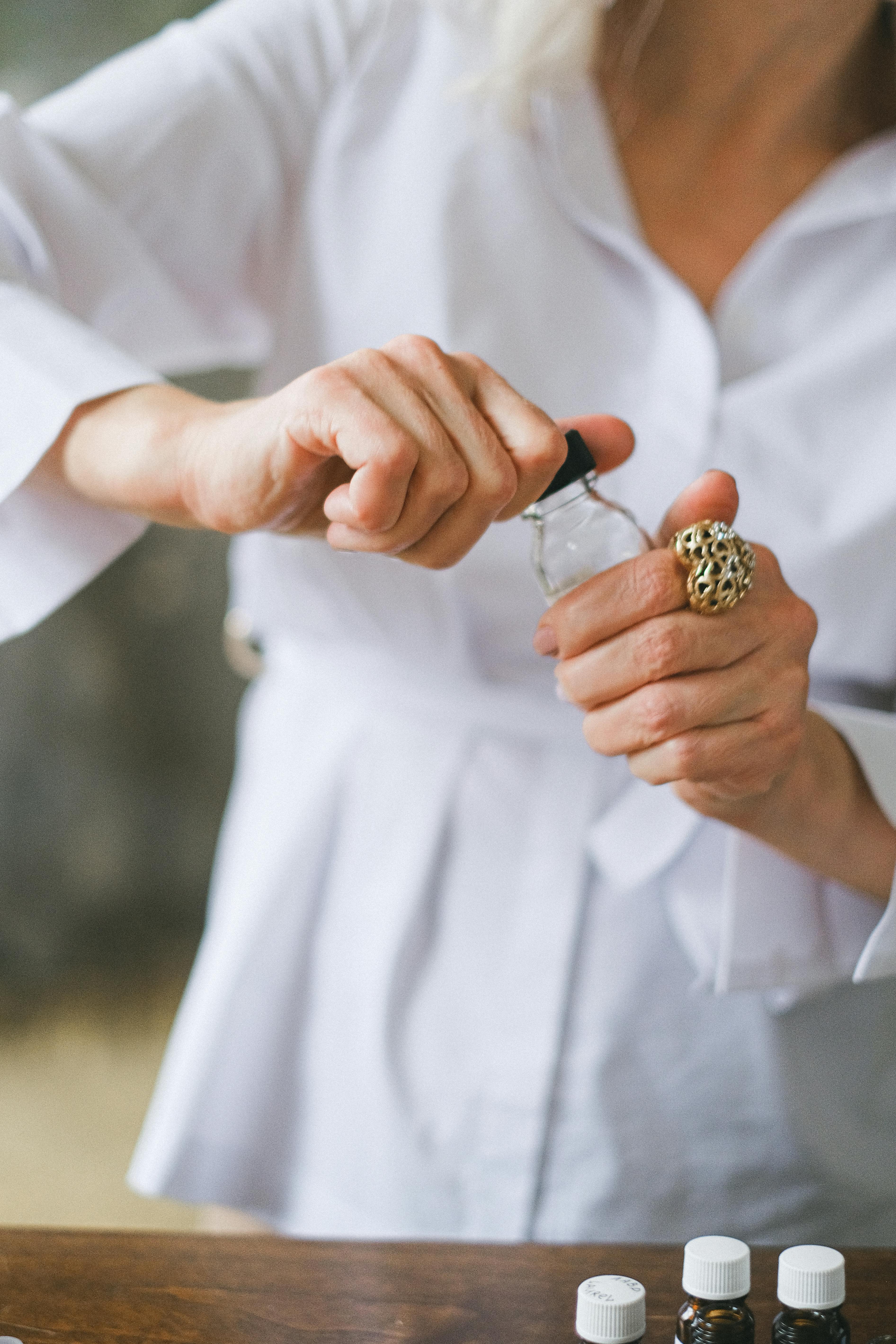 A woman wearing a white shirt opens an essential oil bottle, showcasing hands with jewelry.