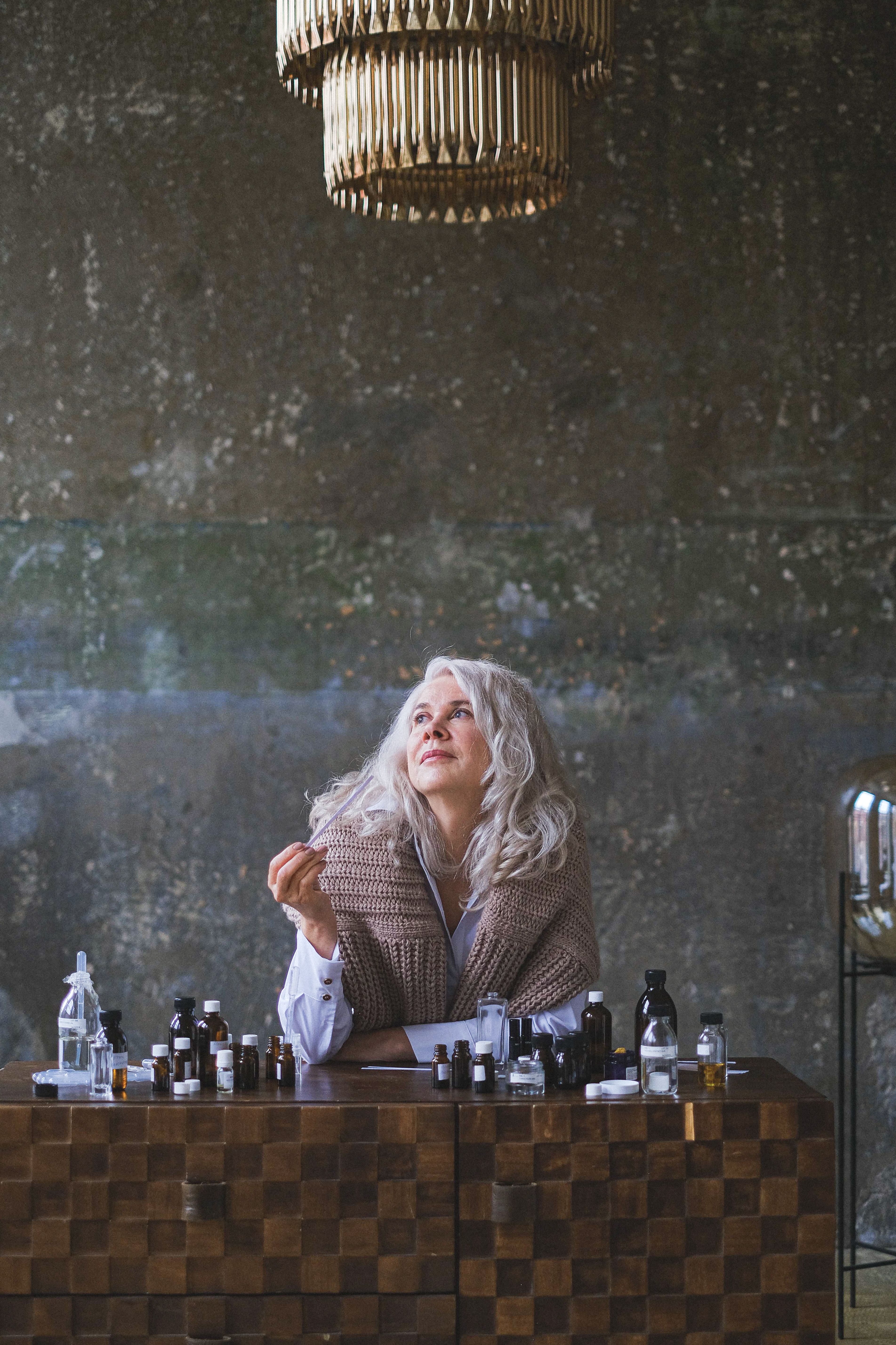Senior woman with gray hair sits at a table with glass bottles, looking upwards and smiling.