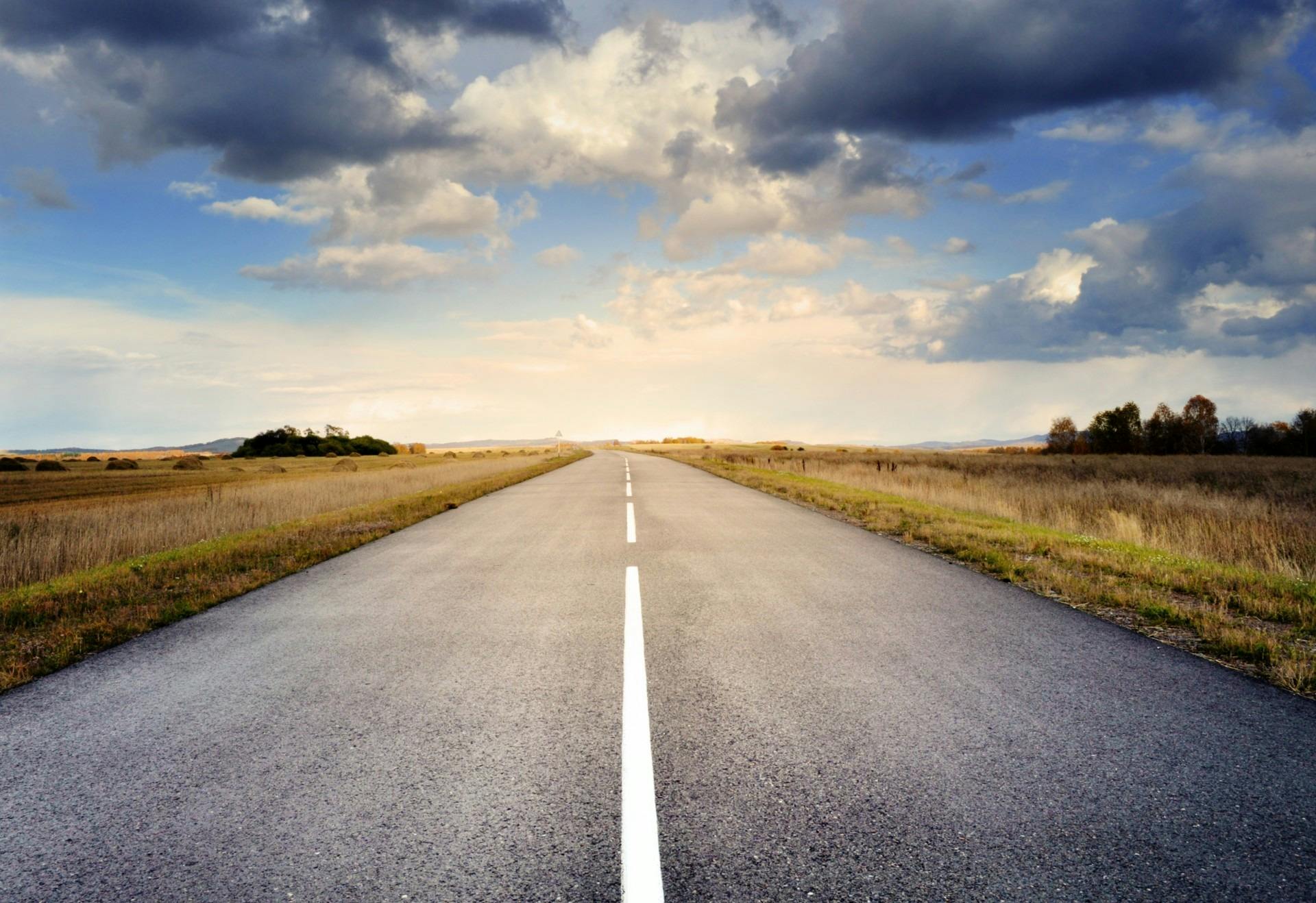 A long straight road stretches through fields under a dramatic cloudy sky.