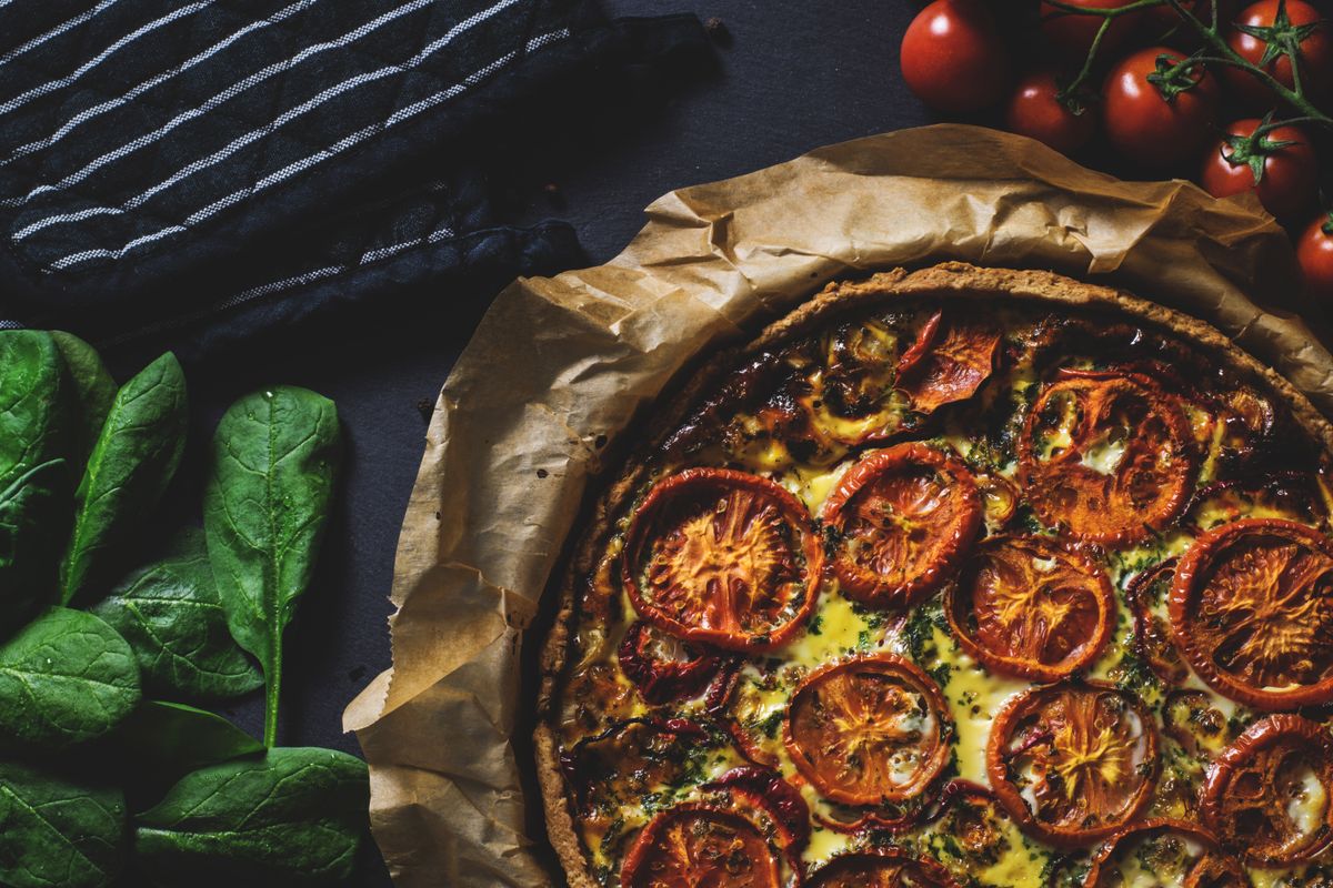 Rustic wholemeal quiche topped with roasted tomato slices on parchment beside spinach leaves