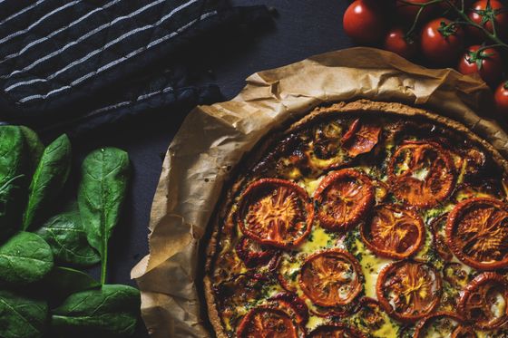 Rustic wholemeal quiche topped with roasted tomato slices on parchment beside spinach leaves