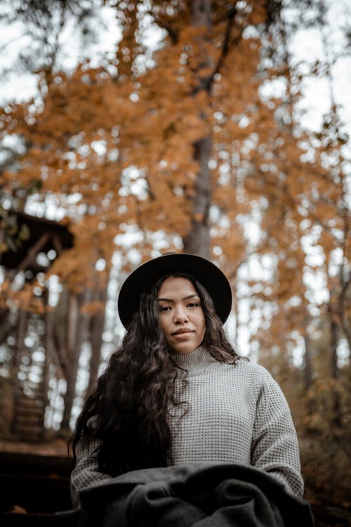 From below charming young ethnic female with long dark hair wearing warm sweater and hat resting on bench against yellow trees in autumn woodland