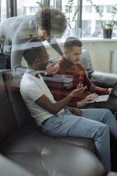 Three men discussing work and technology on a sofa indoors, reflecting teamwork and diversity.
