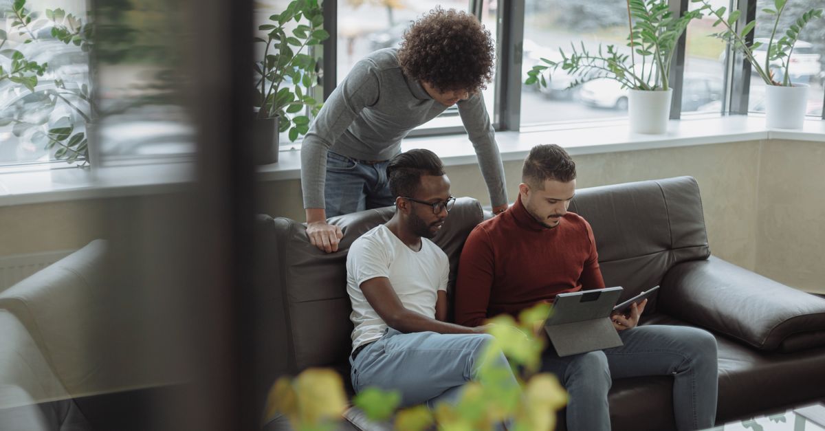 Três homens discutem projetos usando um tablet em um lounge de escritório iluminado, promovendo o trabalho em equipe.