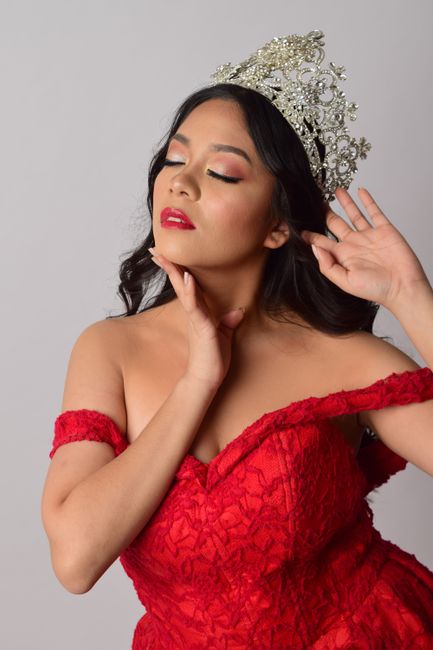 Graceful woman in a red gown with eyes closed, wearing a sparkling tiara against a gray background.