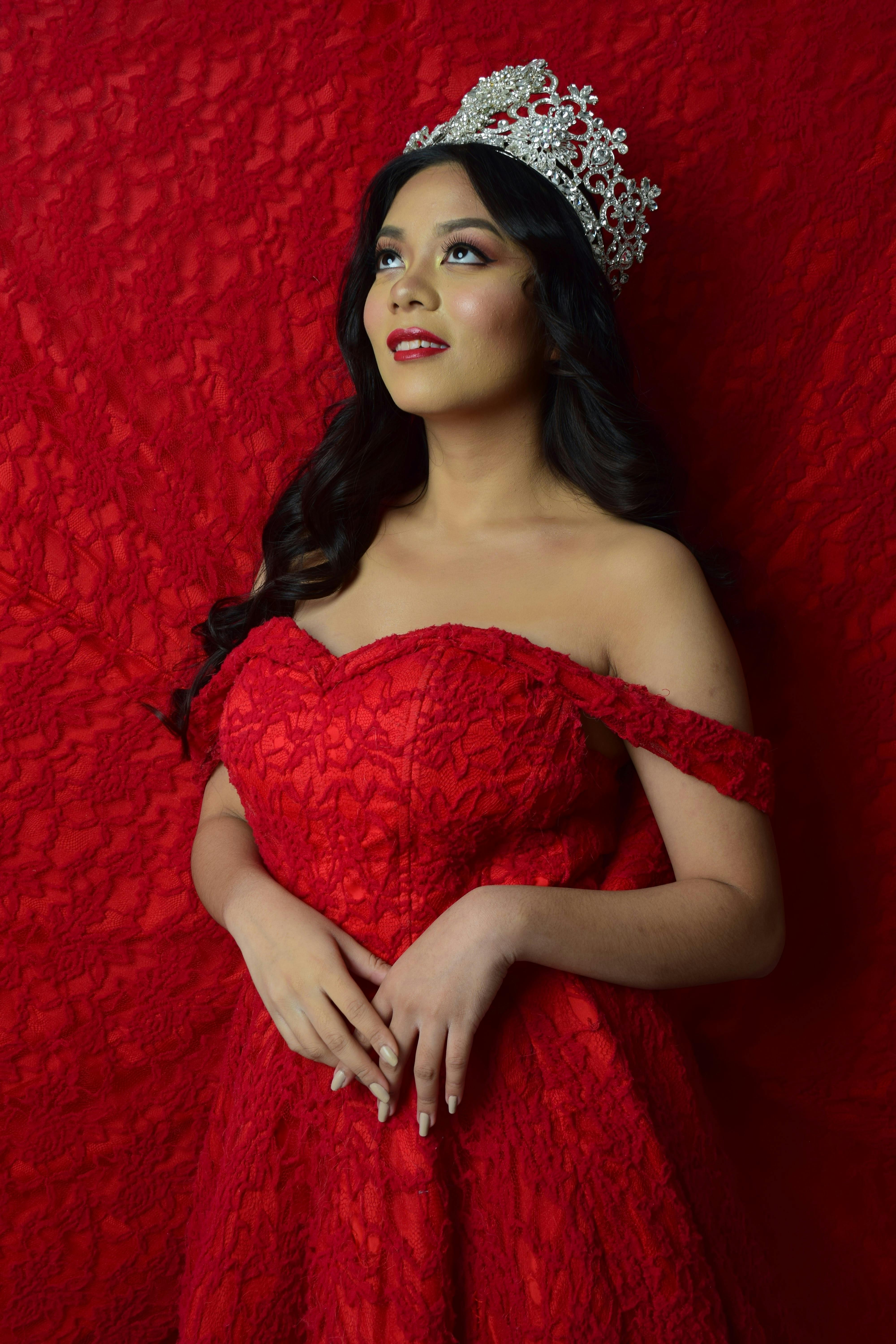 Elegant woman in glamorous red lace gown and crown, exuding sophistication.