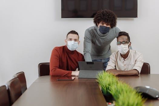 Three colleagues wearing face masks collaborating indoors with a tablet and laptop.