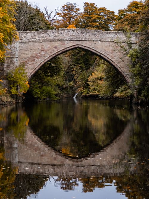 Free A scenic view of a historic stone bridge reflected in a river surrounded by autumn foliage. Stock Photo