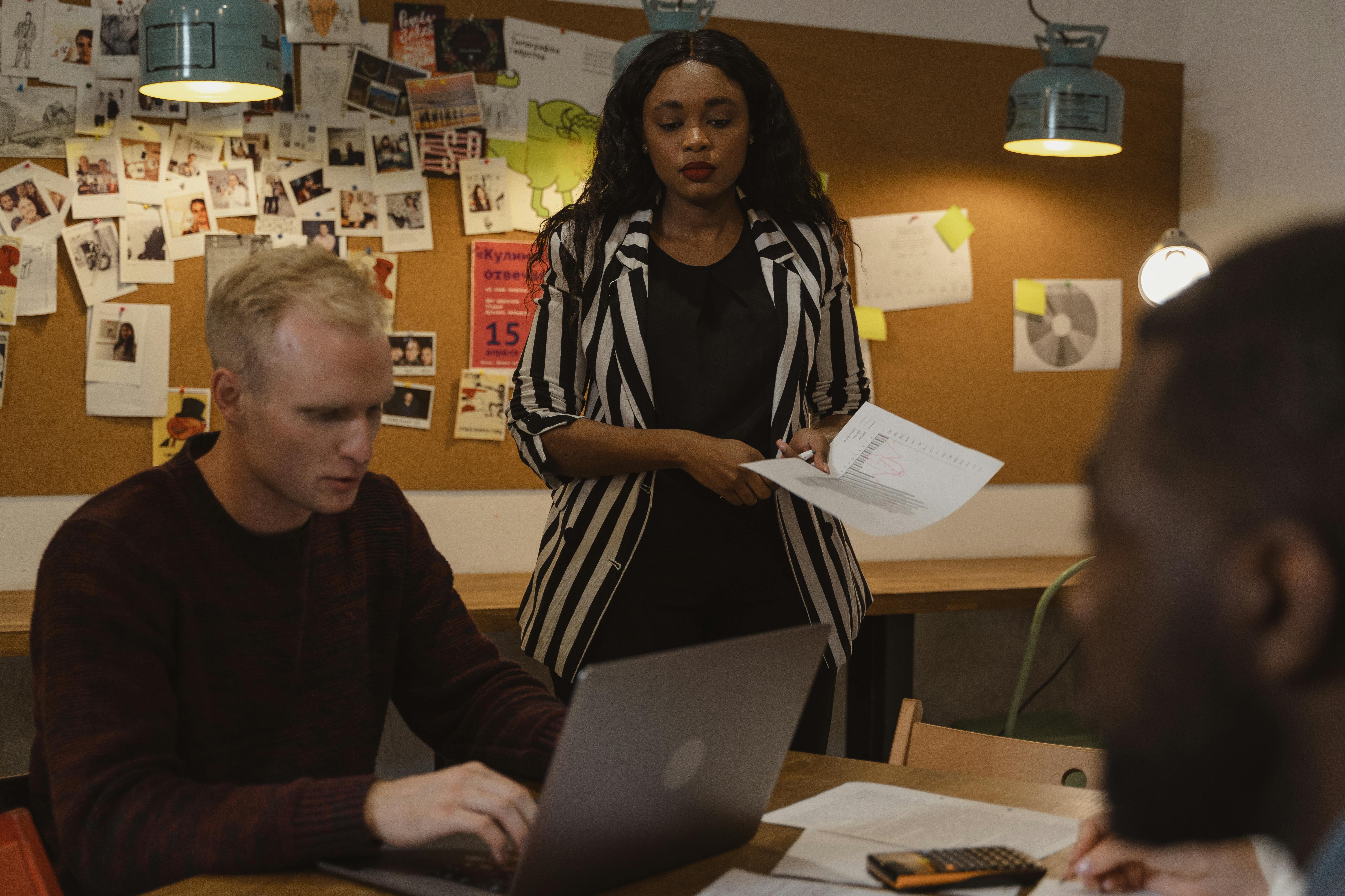 woman in black and white coat standing beside man sitting using laptop