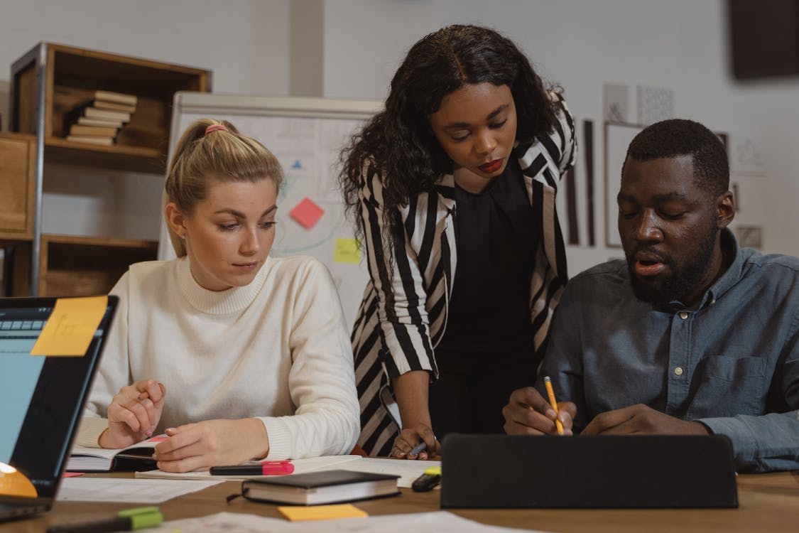 3 people in collaborative group work with a woman in black and white striped shirt leans over between a woman in a white sweater and man in a dark button up shirt.